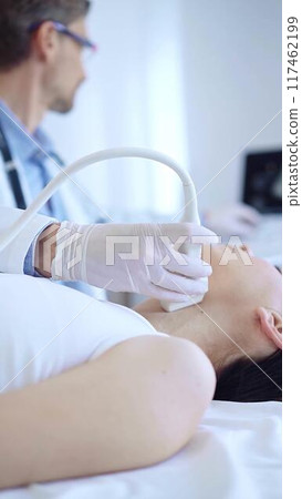 Doctor man wearing white protective gloves is using ultrasound machine to examine neck of female patient in hospital. Medicine, thyroid gland health Doctor man wearing white protective gloves is using ultrasound machine to examine neck of female patient in hospital. Medicine, thyroid gland health 117462199
