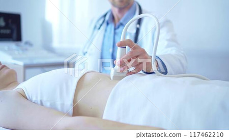 Doctor man wearing a white coat using an ultrasound equipment on a female patient lying down in clinic cabinet. Preventive therapy in medicine Doctor man wearing a white coat using an ultrasound equipment on a female patient lying down in clinic cabinet. Preventive therapy in medicine 117462210