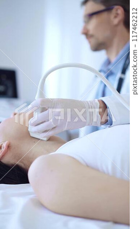 Doctor man wearing white protective gloves is using ultrasound machine to examine neck of female patient in hospital. Medicine, thyroid gland health 117462212