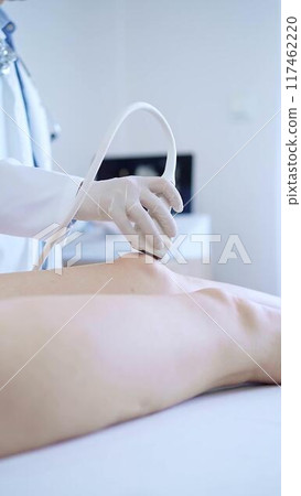 Male doctor with white medical gloves using ultrasound equipment on the knee of a female patient. The results on a monitor in the background. Medicine concept 117462220