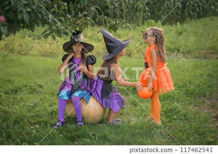 Girls dressed up in witch costumes and child in the form of pumpkin are sitting in garden on Halloween eve Girls dressed up in witch costumes and child in the form of pumpkin are sitting in garden on Halloween eve 117462541