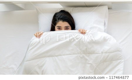A woman is lying in bed, partly hidden under a white blanket, with a playful expression in her eye. 117464877