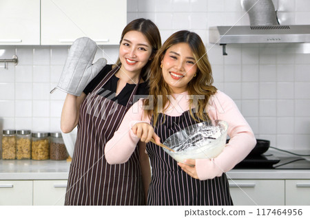 Two women in aprons smiling in the kitchen, one holding an oven mitt, the other mixing batter in a bowl. Two women in aprons smiling in the kitchen, one holding an oven mitt, the other mixing batter in a bowl. 117464956