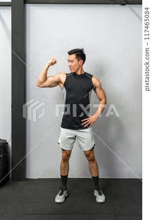 Empowered and strong, a fitness enthusiast male flexes his biceps under the stark, vibrant light of a bustling gym, revealing the result of his perseverance and dedication. 117465084