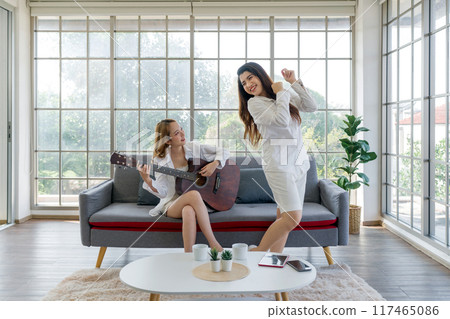Two women are enjoying themselves indoors, one playing guitar and the other dancing, surrounded by a cozy living room setting. Two women are enjoying themselves indoors, one playing guitar and the other dancing, surrounded by a cozy living room setting. 117465086