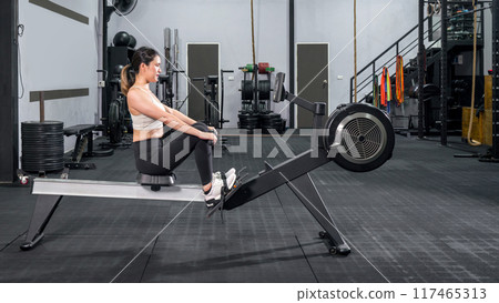 An athletic woman in sportswear is engaged in an intense workout session on a rowing machine inside a well-equipped gym. Health and active lifestyle concept 117465313
