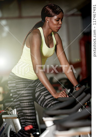 Cycling, gym or black woman on bike machine for cardio training, workout or energy for endurance. Development, exercise or healthy African sports person in club for fitness, challenge or wellness 117467491