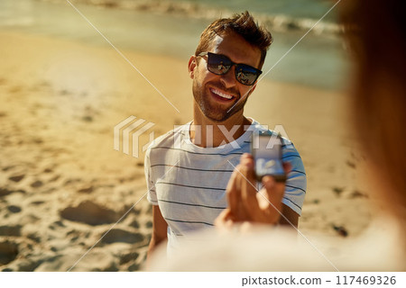 Love, engagement and man on a beach, proposal and relationship with happiness, vacation and support. Smile, ring and people on seaside holiday, sunshine and ask for hand in marriage for romance 117469326
