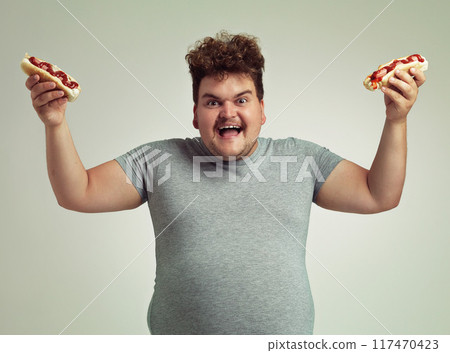 Happy man, fast food and eating hotdog for lunch in studio background for snack, hunger and craving for plus size guy. Male person, hungry and takeaway meal for starving and enjoy while standing Happy man, fast food and eating hotdog for lunch in studio background for snack, hunger and craving for plus size guy. Male person, hungry and takeaway meal for starving and enjoy while standing 117470423