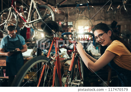 Happy woman, portrait and bicycle mechanic with inspection, fix or maintenance at repair shop. Young female person, cycling technician or engineer working on bike with tools or equipment at workshop 117470579