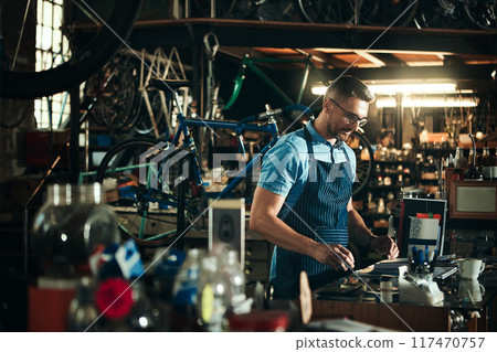 Happy man, bicycle mechanic and workbench with tools in small business for fix, repair or maintenance. Young male person, cycle engineer or bike technician with smile for job or equipment at workshop Happy man, bicycle mechanic and workbench with tools in small business for fix, repair or maintenance. Young male person, cycle engineer or bike technician with smile for job or equipment at workshop 117470757