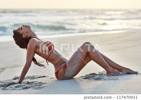 Beach, relax and woman in bikini, sand and rest on ground, nature and trip for vacation in California. Holiday, girl and person in summer with swimsuit, peace and wellness in ocean, calm and sunset 117470931