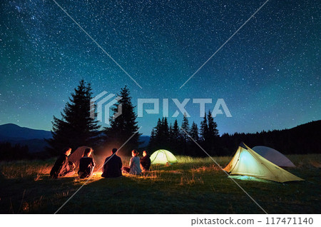 Group of friends hikers resting around campfire, surrounded by illuminated tents under stunning starry sky. Milky Way stretches above, creating magical atmosphere in peaceful mountain wilderness. Group of friends hikers resting around campfire, surrounded by illuminated tents under stunning starry sky. Milky Way stretches above, creating magical atmosphere in peaceful mountain wilderness. 117471140