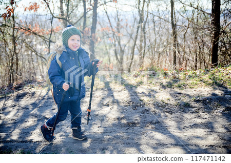 Young child smiling while hiking on forest trail, bundled up in blue winter jacket with fur-lined hood and green striped beanie. Kid holding two trekking poles and enjoying sunny day outdoors. 117471142