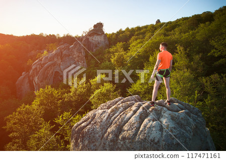 Aerial view of climber standing atop rock formation at Dovbush Rocks in Carpathian mountains, Ukraine. Sun sets, casting warm glow over lush, green forest, distant hills, creating breathtaking view. 117471161