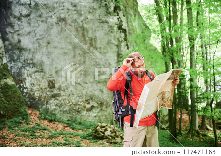 Bearded tourist man looks puzzled with surprised expression, studies map while stands in front of large rock in dense forest. Traveler with backpack and glasses, trying to figure out his direction. 117471163