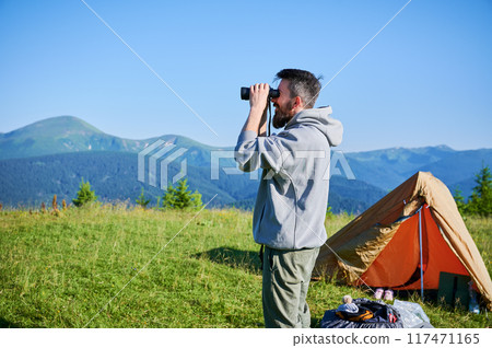 Man stands near orange tent, using binoculars to observe distant mountains. Grassy hilltop, under clear blue sky, lush green forests and rolling hills. Camping gear scattered around tent. 117471165