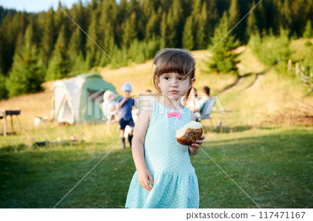 Young girl in light blue dress with pink bow holds bun, standing in grassy camping area. Behind, other children play and adults relax near green tent. Backdrop of lush, pine-covered hills. 117471167