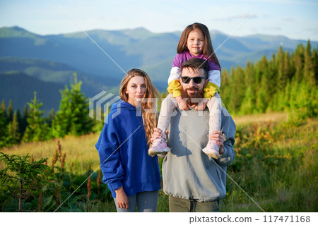 Family stands together in scenic meadow with backdrop of rolling mountains. Father carries his daughter on shoulders, both smiling and enjoying view, while mother stands beside them. 117471168