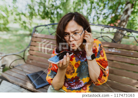 Disgruntled female angrily conversing on her phone on a sunny day in the park. Upset entrepreneur with digital devices, fiercely engaged in a loud phone discussion in an urban green space 117473163