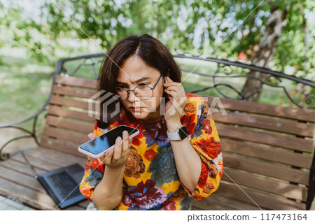 Disgruntled female angrily conversing on her phone on a sunny day in the park. Agitated woman with tech gadgets, furiously making a call amidst in the park 117473164