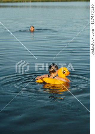 Young girl floating with a yellow duck float at the lake. Her mother is watching over her, ensuring a safe and enjoyable swim Young girl floating with a yellow duck float at the lake. Her mother is watching over her, ensuring a safe and enjoyable swim 117473180