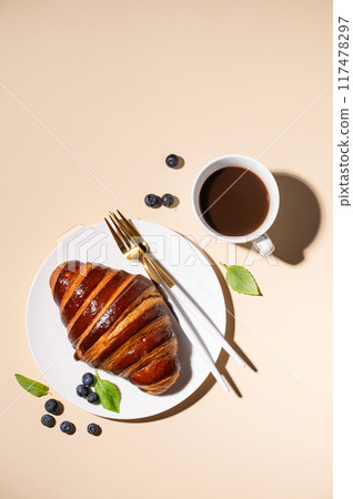 Freshly baked croissant with chocolate on a white plate and blueberries on a yellow background  117478297