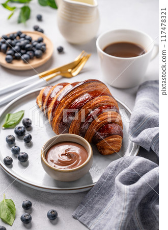 Fresh croissant with chocolate on a plate on a light background with a blueberries, cup of coffee 117478321