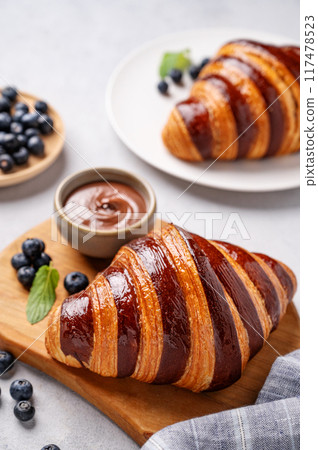 Fresh croissants with chocolate on a cutting board on a light background with a blueberries close up 117478523