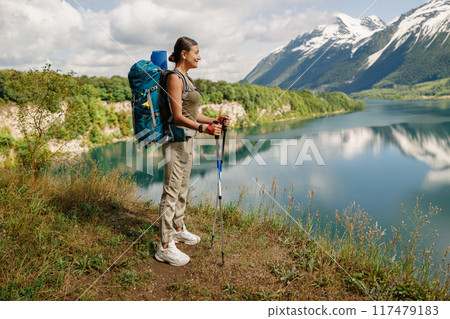 A Woman Enjoys Hiking Beside a Serene Lake While Majestic Mountains Rise in the Background A Woman Enjoys Hiking Beside a Serene Lake While Majestic Mountains Rise in the Background 117479183
