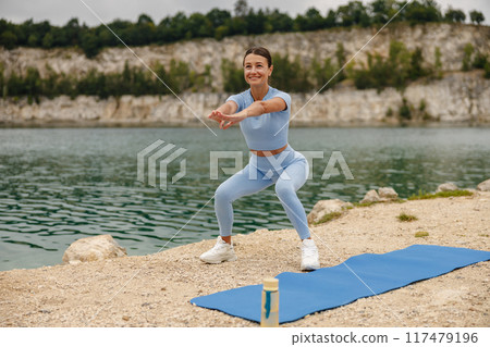 A dedicated woman is exercising outdoors, doing squats beside a beautiful body of water 117479196
