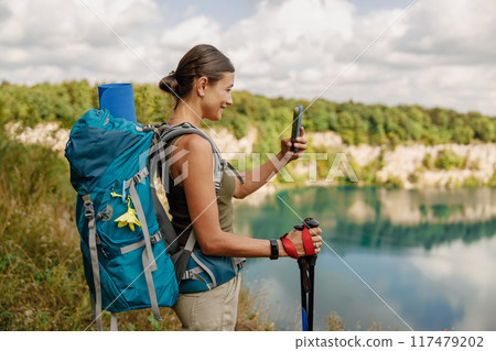 A Hiker is Enjoying a Relaxing Break at a Scenic Lake While Using a Smartphone to Capture the Moment 117479202