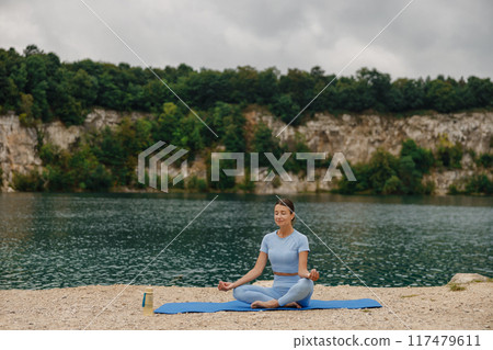 A woman deeply meditating by a serene lake, surrounded by the beauty of nature and tranquility 117479611