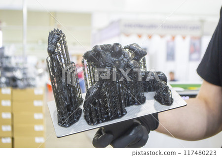 Black model with supports printed on 3D printer close-up. Man holding in hand object created on 3D printer from hardened rubber under influence of ultraviolet light. SLA 3D printing. Stereolithography 117480223