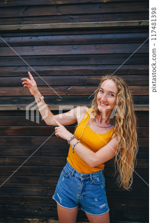 Young woman in yellow tank top points excitedly at a wooden wall in summer 117481398