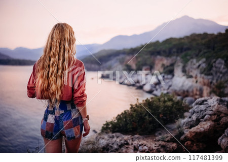 Woman gazes at sunset over a coastal landscape near mountains at dusk 117481399