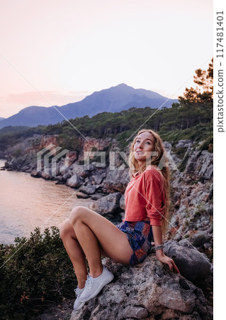 Young woman sitting on a rock by the coastline at sunset, with mountains in the background 117481401