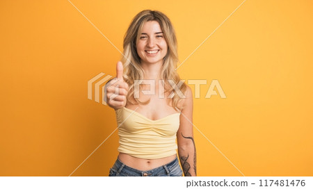 Happy young woman showing thumb up gesture on yellow background Happy young woman showing thumb up gesture on yellow background 117481476