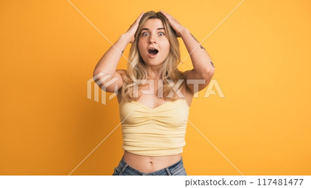 Smiling young woman standing with widened eyes on yellow background 117481477