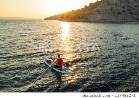 Man wearing an orange vest navigates his paddleboard through gentle waves, with the warm glow of the setting sun Man wearing an orange vest navigates his paddleboard through gentle waves, with the warm glow of the setting sun 117482271