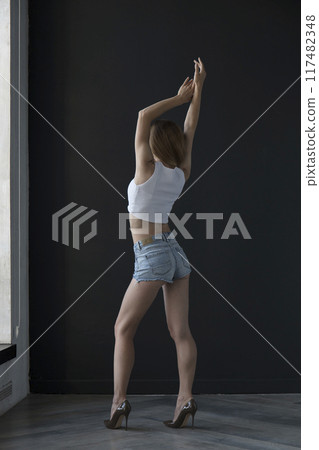 young woman in a white tank top and denim shorts on a dark background, full length portrait 117482348