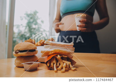 To eat or not. Cheerless plump woman looking at the burger while thinking to eat it or not To eat or not. Cheerless plump woman looking at the burger while thinking to eat it or not 117484467