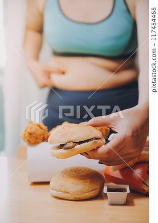 To eat or not. Cheerless plump woman looking at the burger while thinking to eat it or not To eat or not. Cheerless plump woman looking at the burger while thinking to eat it or not 117484468