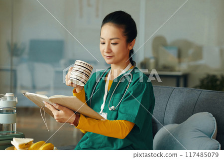 Nurse Reading Book and Holding Coffee Cup in Hospital Office Nurse Reading Book and Holding Coffee Cup in Hospital Office 117485079