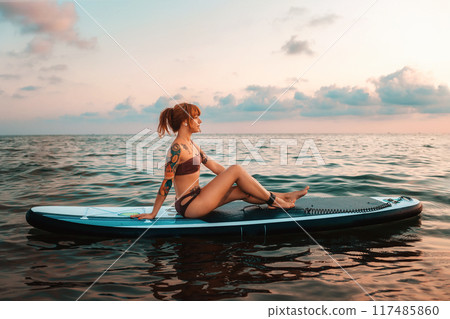A tanned young woman with a tattoo poses sitting on a sup board swimming at water. Copy space. Sunset sky. The concept of sports recreation and surfing 117485860