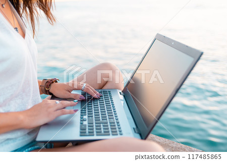 A woman works remotely on a laptop. Hands close-up. Sea in the background. The concept of freelancing 117485865