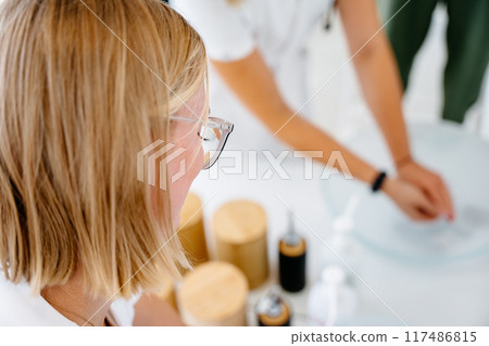 A woman with glasses is looking at a table with various items on it. The table has a glass of water, a bottle, and a cup 117486815