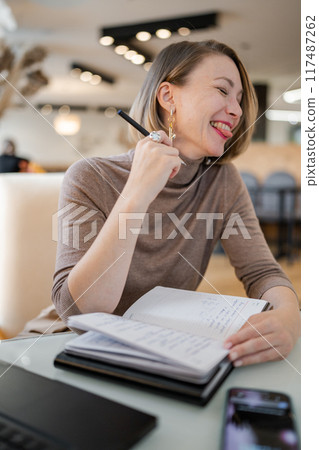 Young laughing woman working on laptop in cozy cafe with other people engaging in conversation in background. Concentrated study in co-working space. Education and resource searching concept Young laughing woman working on laptop in cozy cafe with other people engaging in conversation in background. Concentrated study in co-working space. Education and resource searching concept 117487262