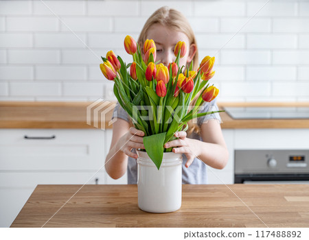 The girl puts a beautiful bouquet of red and yellow tulips in a white vase on a wooden countertop The girl puts a beautiful bouquet of red and yellow tulips in a white vase on a wooden countertop 117488892