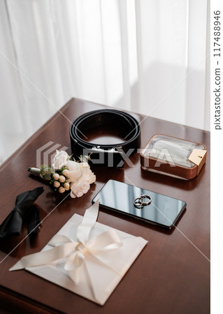 Groom's wedding essentials on table, featuring rings on phone, bow tie, boutonniere, perfume, belt 117488946